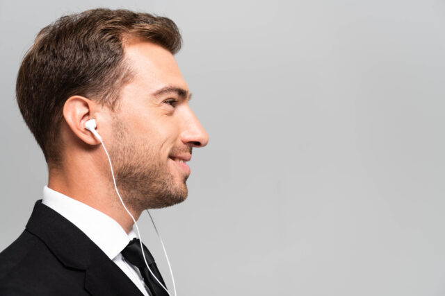 side view CARATTERISTICHE DELL'IMPRENDITORE DIGITALE: SAPER ASCOLTARE handsome and smiling businessman in suit listening music with earphones isolated on Imprenditore ascolto